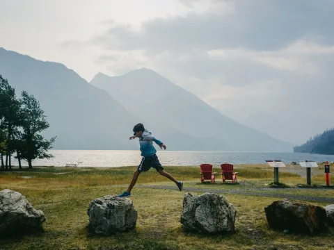 Child running across a lakeside lawn with mountains and red chairs in the distance