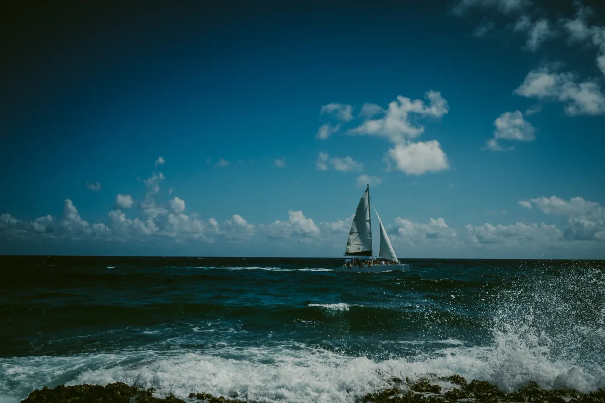 Small sailboat crossing bright blue ocean water under scattered clouds