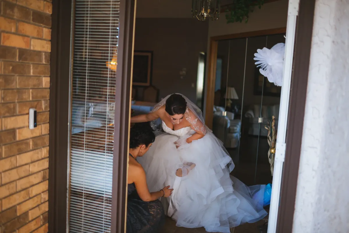 View through an open doorway of a bride in a tulle ballgown being helped with her dress