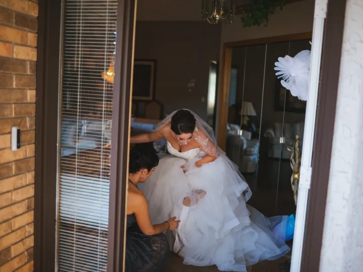 View through an open doorway of a bride in a tulle ballgown being helped with her dress