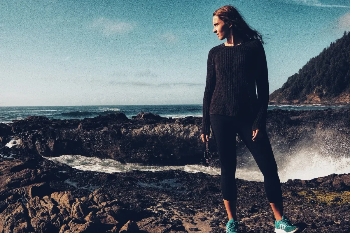 Woman standing on a rocky shore in dark clothing with ocean behind her