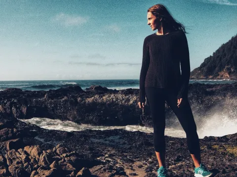 Woman standing on a rocky shore in dark clothing with ocean behind her