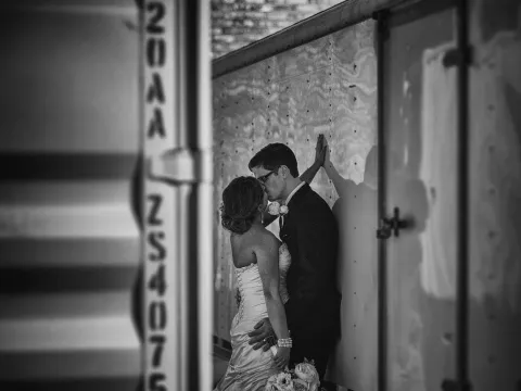 Black and white photo of bride and groom kissing between industrial metal structures