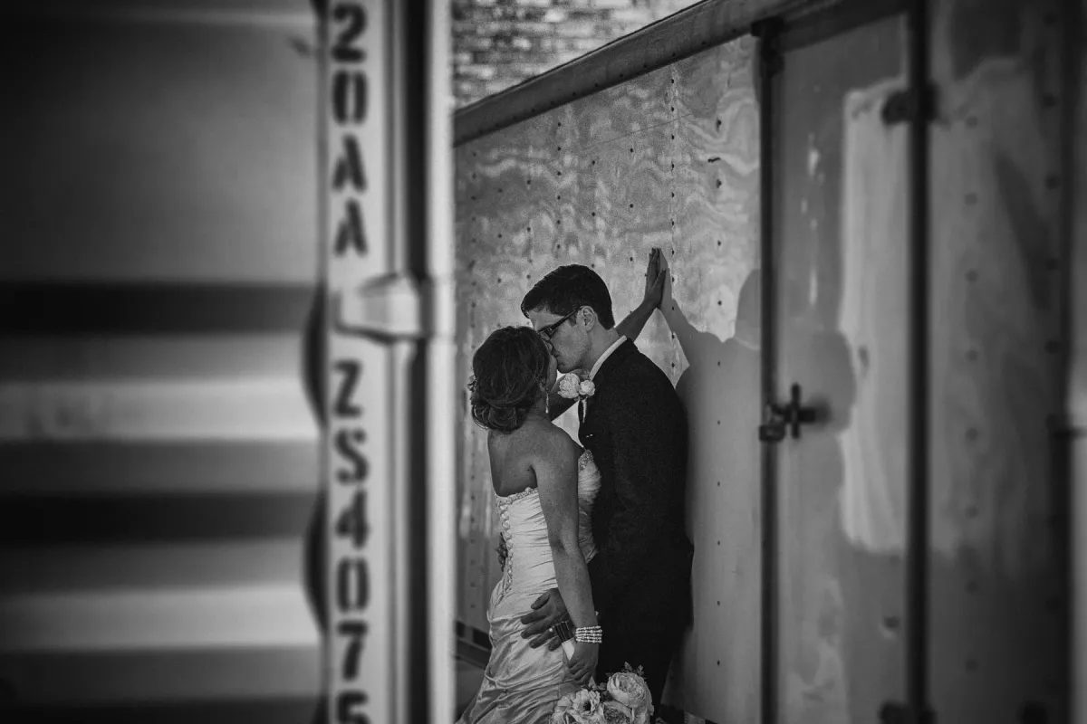 Black and white photo of bride and groom kissing between industrial metal structures