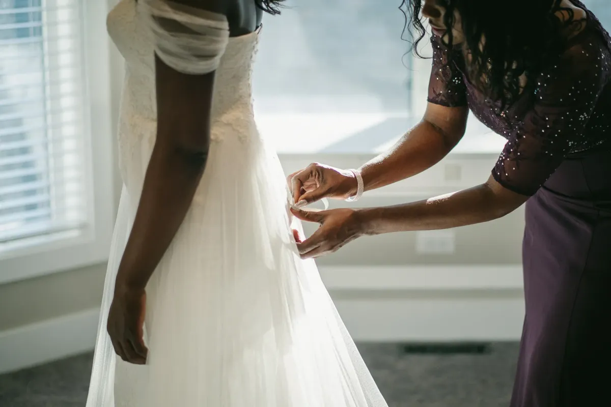 Close-up of hands fastening the back of a bride's lace dress beside a sunny window