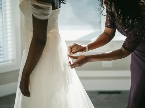 Close-up of hands fastening the back of a bride's lace dress beside a sunny window