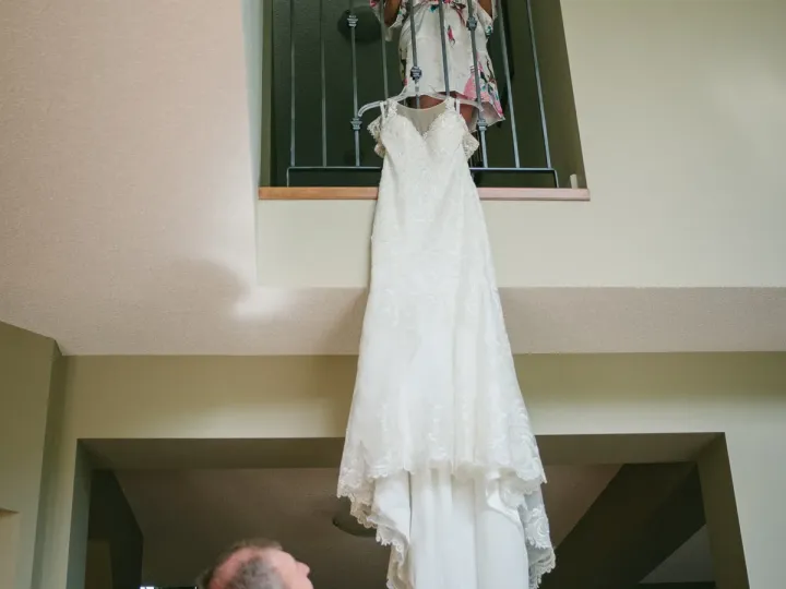 Bride on an upper balcony looking down at her hanging wedding dress while her father looks up below