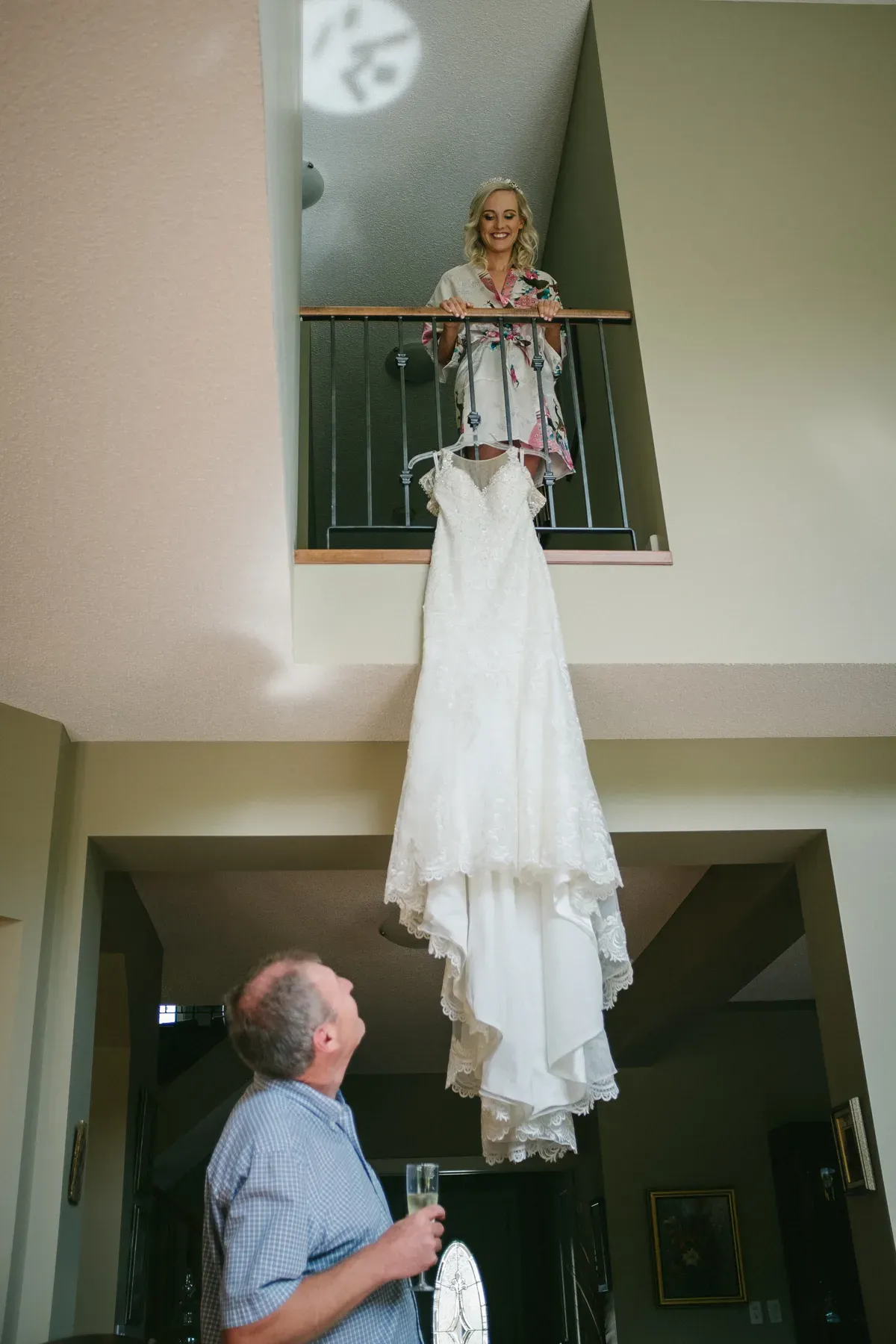 Bride on an upper balcony looking down at her hanging wedding dress while her father looks up below