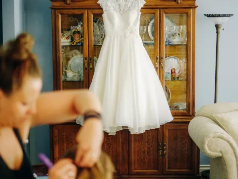 White tea-length wedding dress hanging on a wooden china cabinet with blurred hair styling in foreground