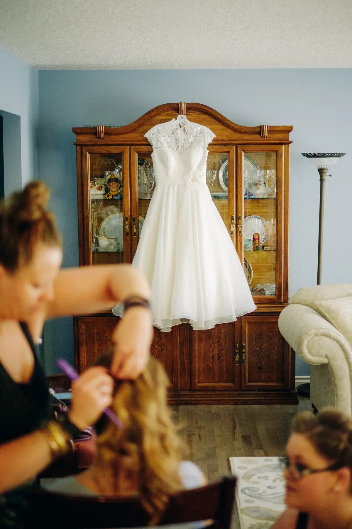White tea-length wedding dress hanging on a wooden china cabinet with blurred hair styling in foreground
