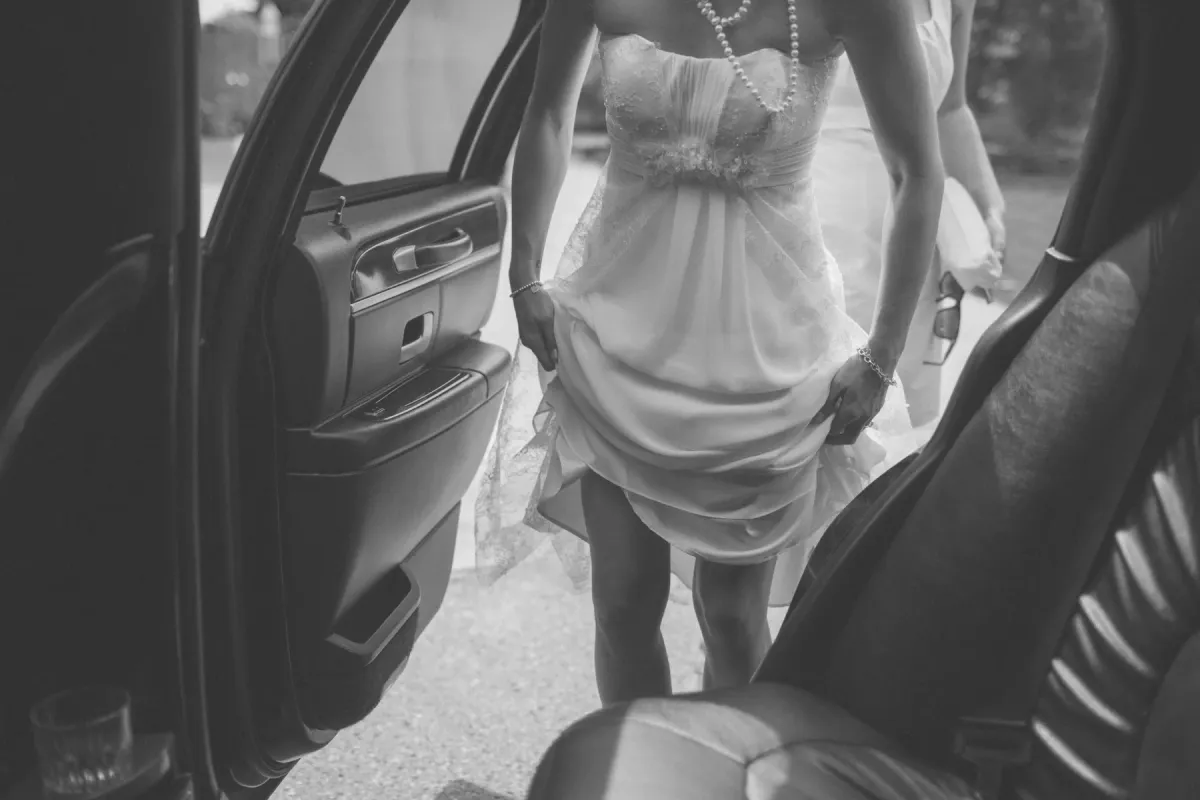 Black and white view from a car of a bride lifting her dress as she steps out