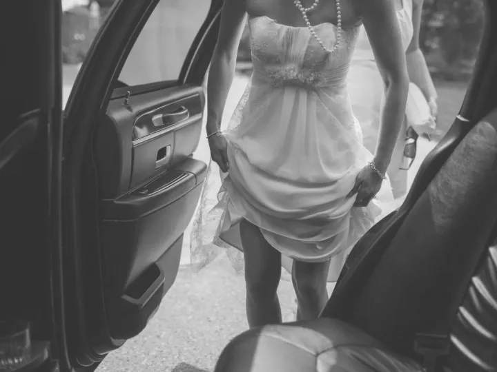 Black and white view from a car of a bride lifting her dress as she steps out
