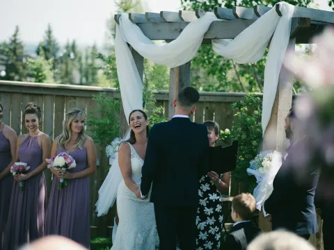 Bride laughing during an outdoor ceremony under a wooden arbor with mauve bridesmaids nearby