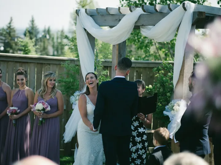 Bride laughing during an outdoor ceremony under a wooden arbor with mauve bridesmaids nearby
