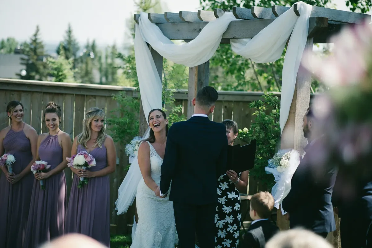 Bride laughing during an outdoor ceremony under a wooden arbor with mauve bridesmaids nearby