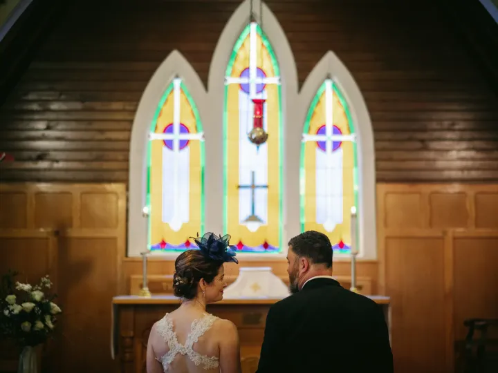 Bride and groom from behind at a wooden church altar before stained glass windows