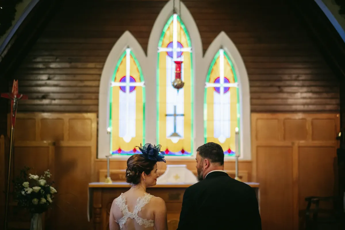 Bride and groom from behind at a wooden church altar before stained glass windows