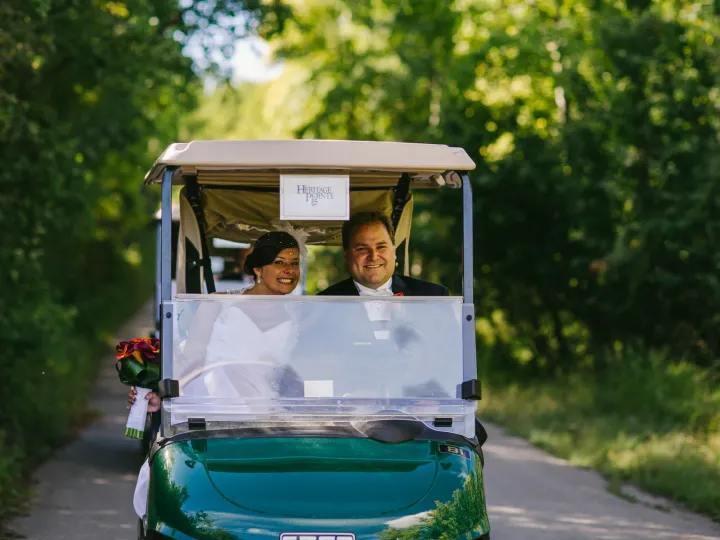 Bride and groom smiling in a green Heritage Pointe golf cart on a tree-lined path