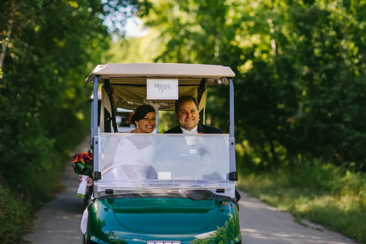 Bride and groom smiling in a green Heritage Pointe golf cart on a tree-lined path