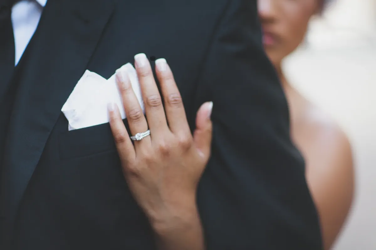 Close-up of a bride's hand with a diamond ring resting on a groom's black tuxedo lapel