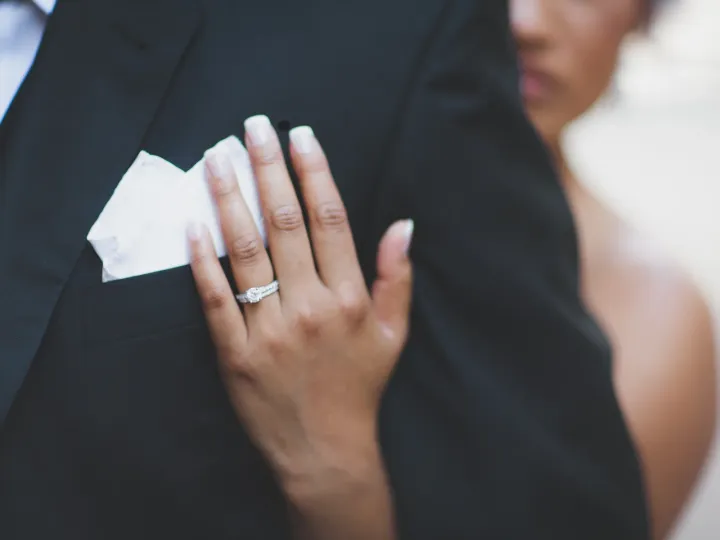 Close-up of a bride's hand with a diamond ring resting on a groom's black tuxedo lapel