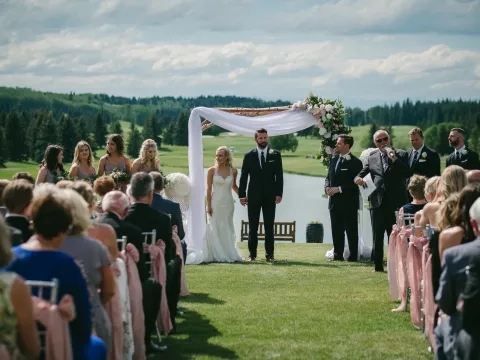 Wide outdoor wedding ceremony with floral wooden arch, dusty rose chairs, and Alberta hills beyond