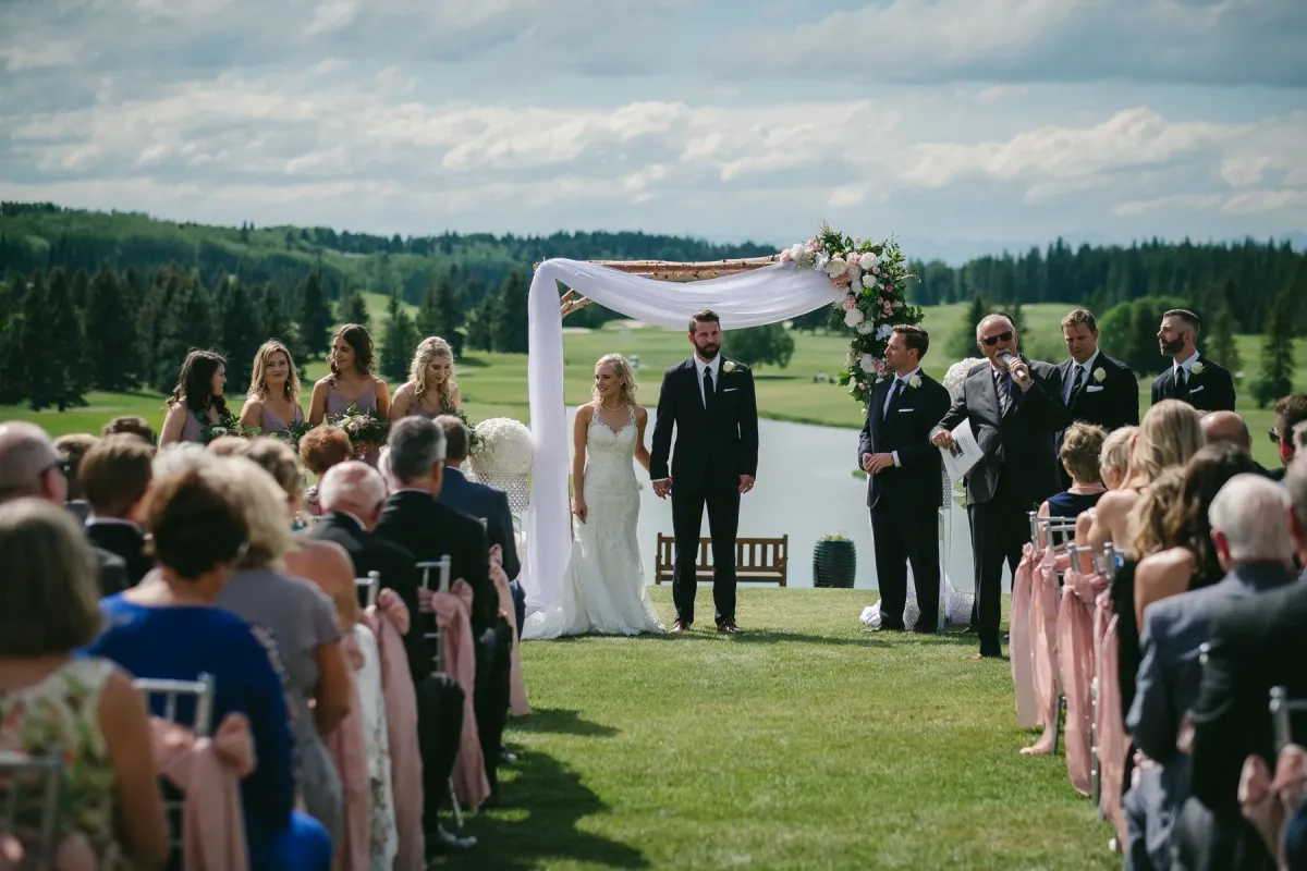 Wide outdoor wedding ceremony with floral wooden arch, dusty rose chairs, and Alberta hills beyond