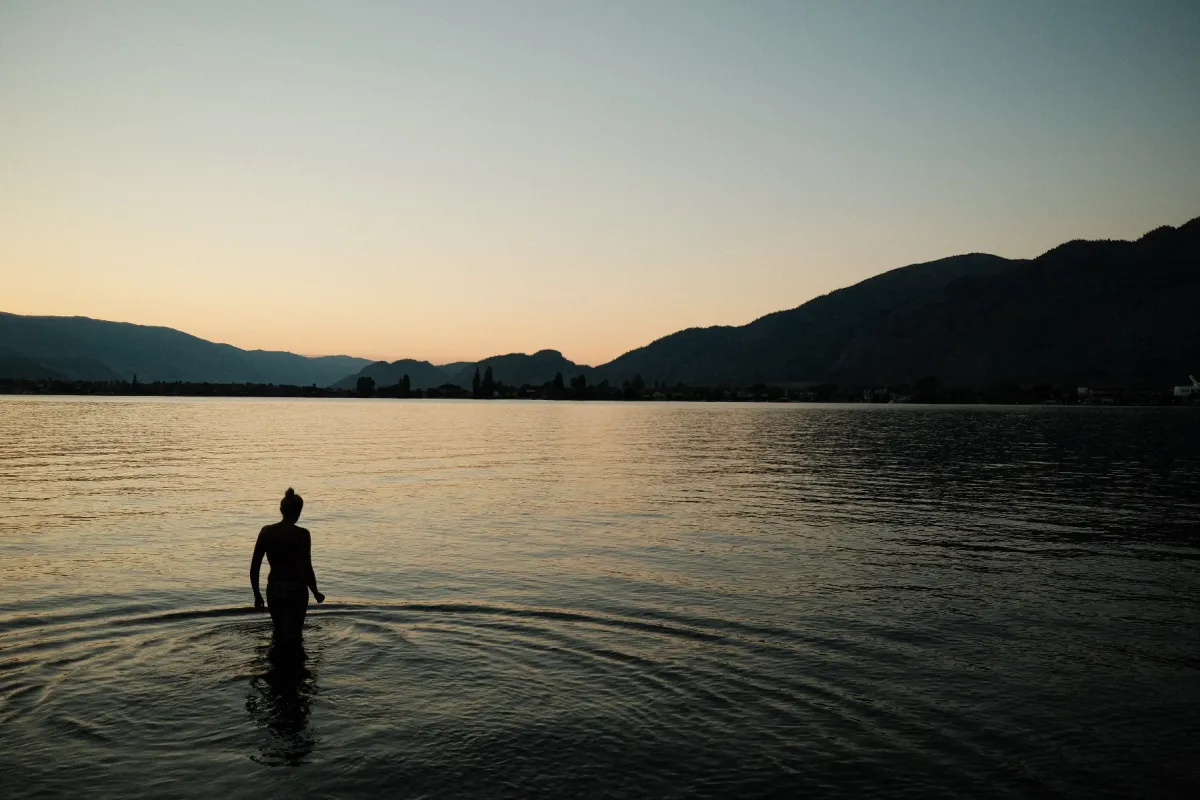 Person standing in calm lake water at dusk between dark mountain shores