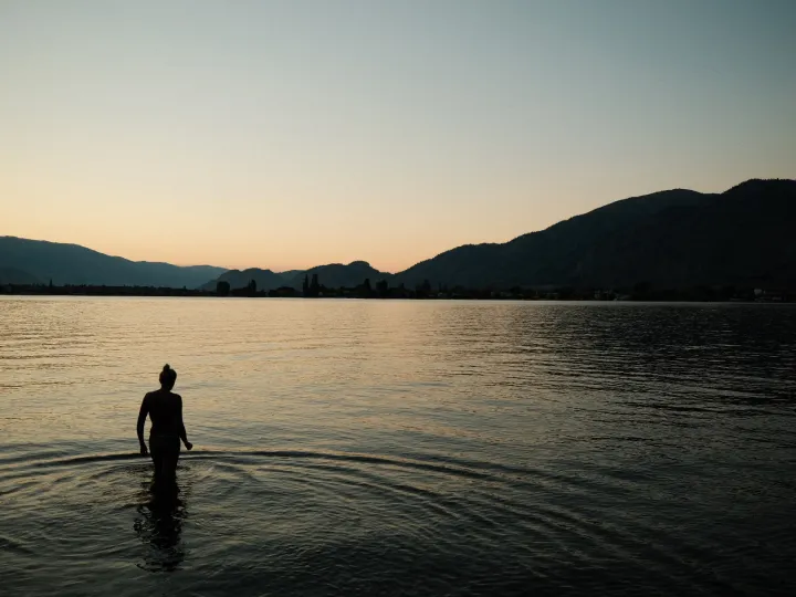 Person standing in calm lake water at dusk between dark mountain shores