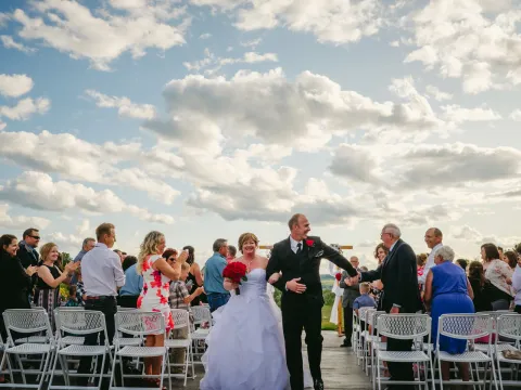 Newlyweds walking down an outdoor aisle on a wooden deck as guests cheer under a cloudy sky