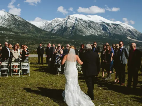 Bride walking down a grassy outdoor aisle with guests and snow-capped mountains behind