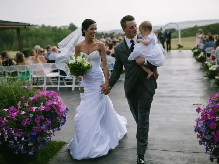 Bride and groom walking an outdoor aisle while the groom carries a toddler in a white dress
