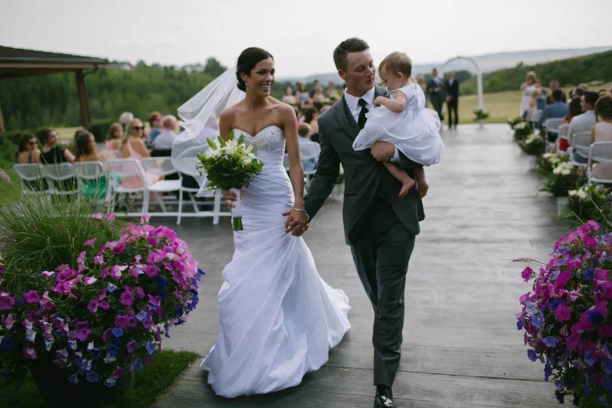 Bride and groom walking an outdoor aisle while the groom carries a toddler in a white dress