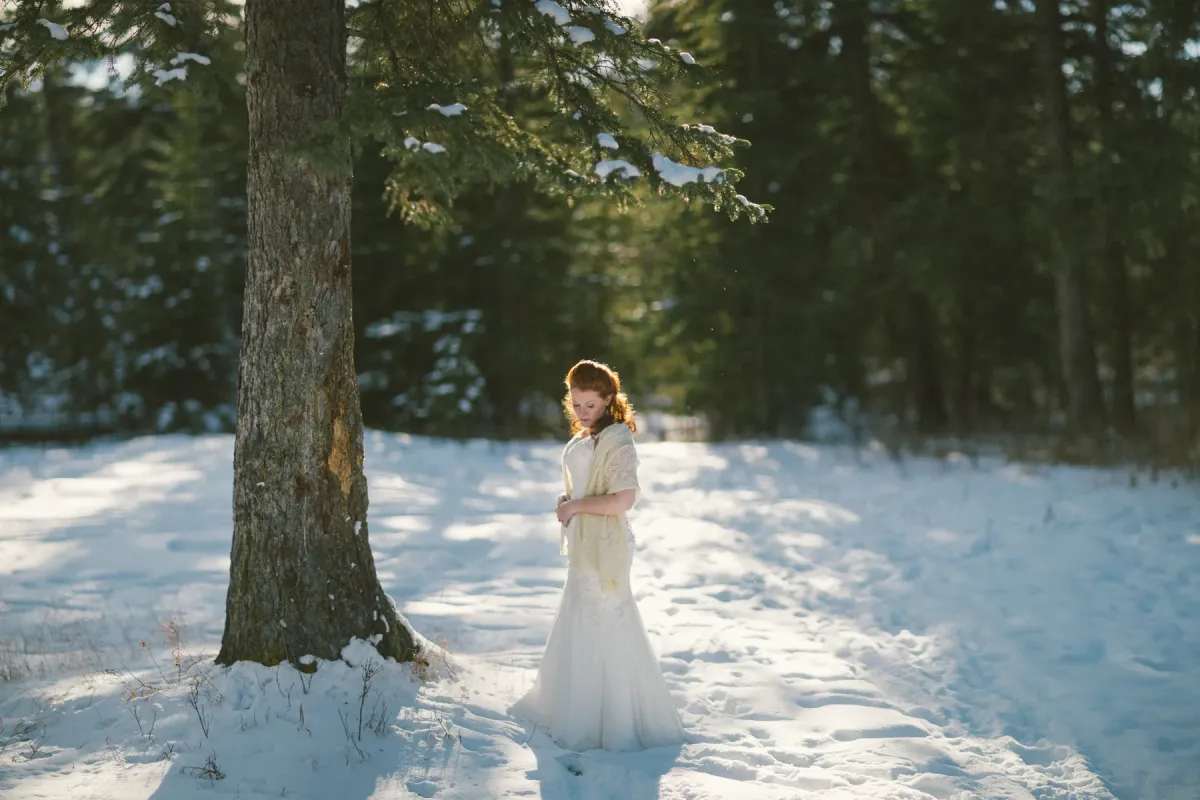 Red-haired bride in a white gown and cream shawl standing in a snowy evergreen forest