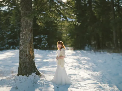 Red-haired bride in a white gown and cream shawl standing in a snowy evergreen forest