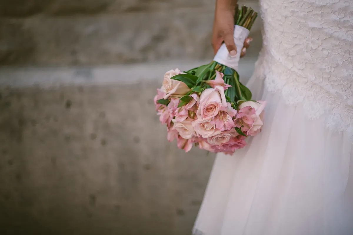 Close-up of a bridal bouquet of pale pink roses held beside a white lace wedding dress