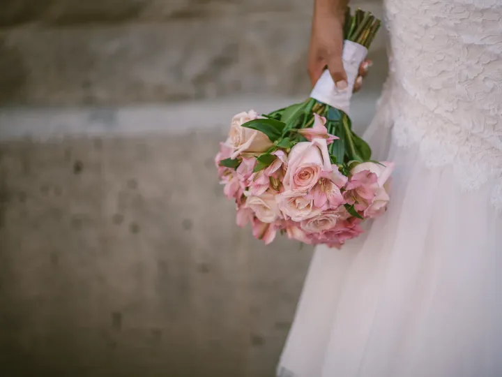 Close-up of a bridal bouquet of pale pink roses held beside a white lace wedding dress