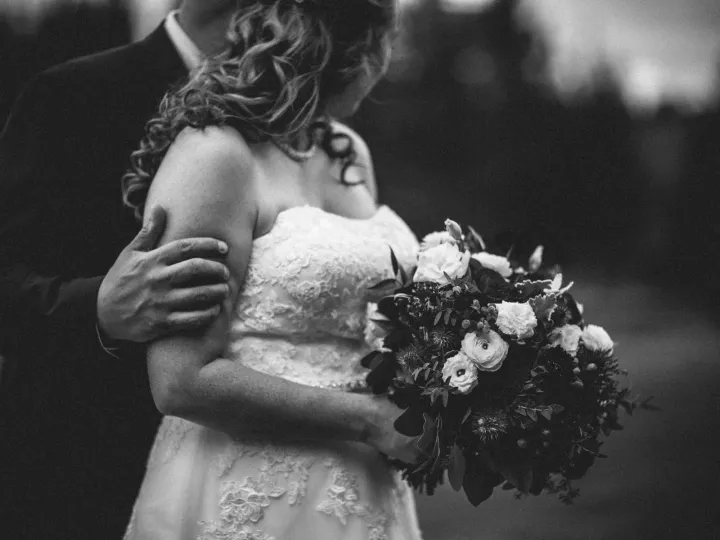Black and white close-up of a groom's hand on a bride's arm as she holds a floral bouquet