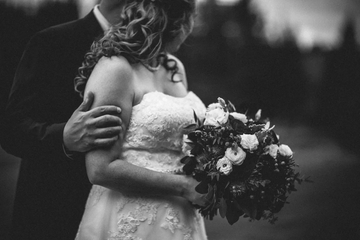 Black and white close-up of a groom's hand on a bride's arm as she holds a floral bouquet