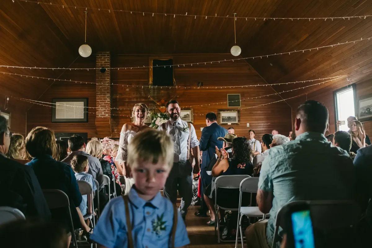 Bride and groom laughing while walking down a wood-paneled aisle with a young boy ahead