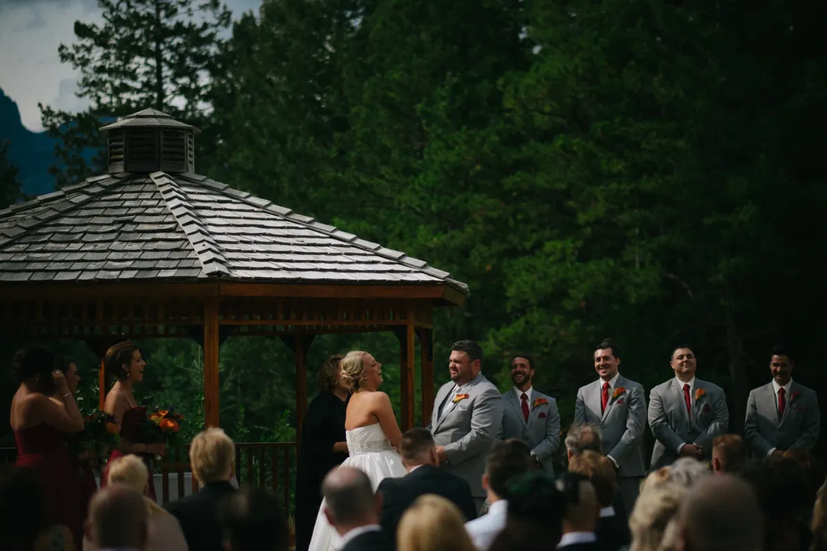 Bride and groom laughing during a ceremony under a wooden gazebo with evergreen forest behind