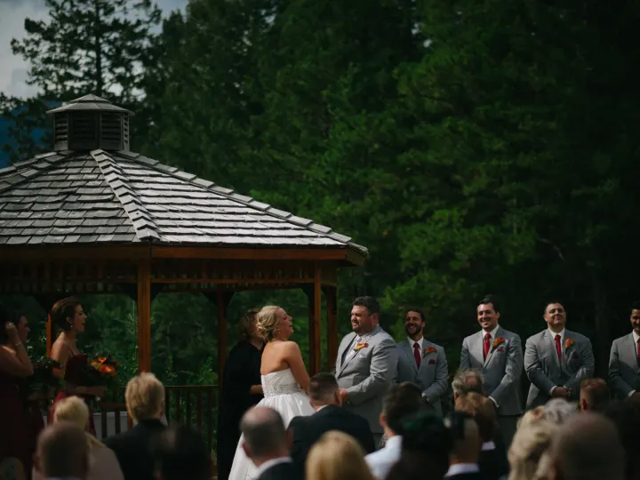 Bride and groom laughing during a ceremony under a wooden gazebo with evergreen forest behind