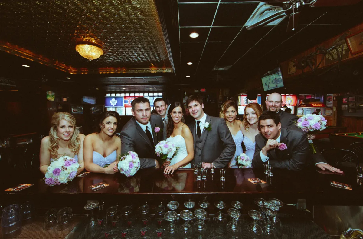 Bride, groom, and wedding party smiling behind a wooden pub bar with shot glasses