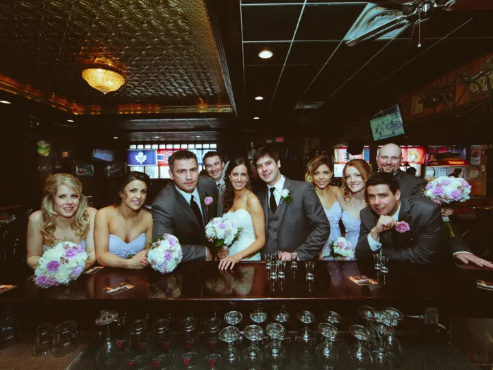 Bride, groom, and wedding party smiling behind a wooden pub bar with shot glasses