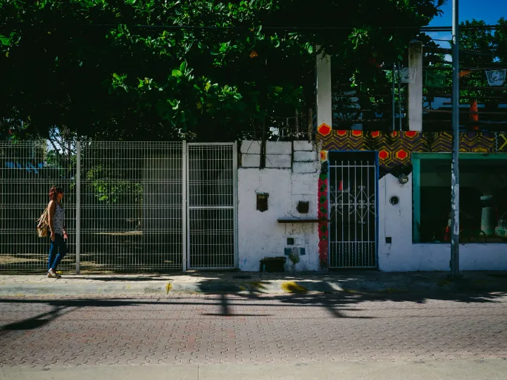 Person walking past a white wall and gate in strong afternoon sunlight